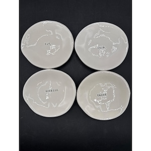 May include: Four off-white ceramic bowls with irregular edges. Each bowl has a word in black text: "EAT," "CHOW," "NOURISH," and "ENJOY." The bowls are arranged in a square on a dark surface.