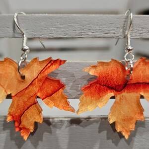 May include: A pair of earrings featuring a resin leaf design with a red, orange, and yellow color scheme. The leaves are attached to silver hooks.