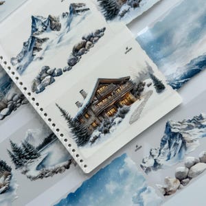 May include: A collection of stickers featuring watercolor-style winter landscapes, including a multi-story cabin with warm glowing windows, snow-covered pine trees, rocky mountain peaks, and blue sky scenes, all printed on a white backing sheet with holes.