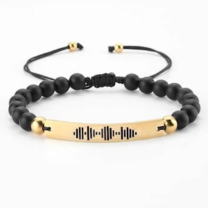以下が含まれることがあります： 黒いビーズブレスレット。金色のバーには音波のデザインが施されています。ブレスレットは黒い調節可能なコードと金色のアクセントビーズが付いています。音波のデザインは黒です。