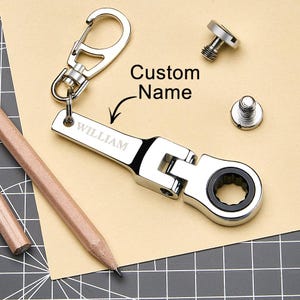 May include: A silver metal wrench keychain with the name "WILLIAM" engraved on it. The keychain has a carabiner clip and a pivoting head. The words "Custom Name" are also visible.