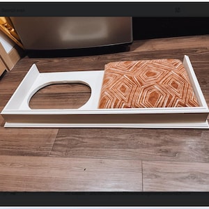 Puede incluir: Cama de madera blanca para mascotas con un cojín extraíble. El cojín tiene un patrón geométrico en tonos naranja y blanco.