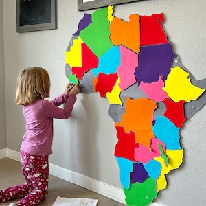 May include: A young child is putting together a colorful puzzle map of Africa. The puzzle pieces are made of foam and are brightly colored. The child is wearing a pink shirt and red pants.