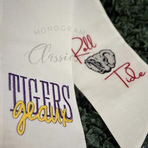 May include: Two white fabric towels with embroidered designs. One towel has the words "Tigers Geaux" in purple and gold thread. The other towel has the words "Roll Tide" in red thread with an elephant mascot in purple thread.