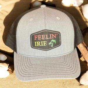 May include: A gray and black trucker hat with a black leather patch that says "Feelin' Irie" in red, yellow, and green lettering. The patch also features two green palm trees.