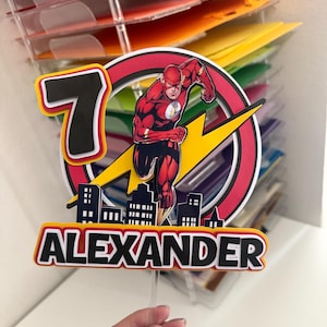 May include: A birthday cake topper featuring The Flash superhero. The design includes a red and black circular frame, a yellow lightning bolt, the number 7, and the name "ALEXANDER" in bold black letters. The Flash is depicted in his signature red suit.