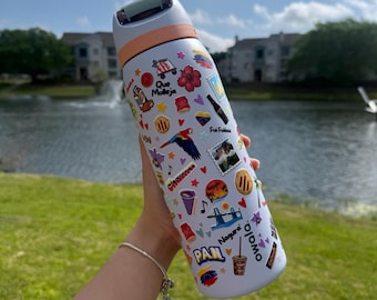 May include: A light blue Owala water bottle covered in colorful stickers, including a parrot, flowers, and the word "PAN." The bottle has a light orange band and a green top. The background shows a lake and buildings.