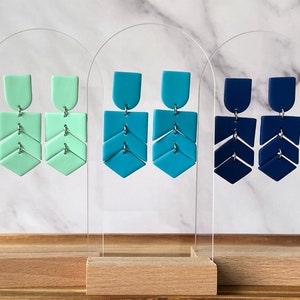 May include: Three pairs of geometric statement earrings displayed on a white acrylic stand. The earrings are made of polymer clay and feature a chevron design. The earrings are available in three colors: mint green, teal blue, and navy blue.