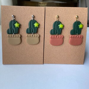 May include: Two pairs of green cactus earrings with yellow flowers. The earrings are made of polymer clay and have a brown pot. One pair has silver ear wires and the other pair has gold ear wires.