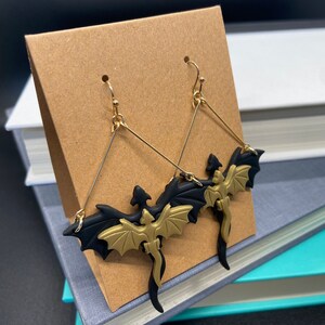 May include: A pair of gold and black dragon earrings. The dragons are made of polymer clay and have a detailed design. The earrings are hanging from gold hooks.