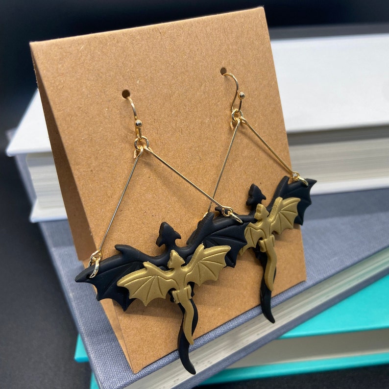May include: A pair of gold and black dragon earrings. The dragons are made of polymer clay and have a detailed design. The earrings are hanging from gold hooks.