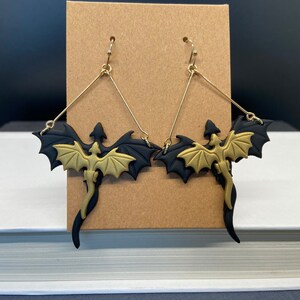 May include: A pair of gold and black dragon earrings. The dragons are in a flying pose with their wings spread out. The earrings are made of polymer clay and have gold hooks.