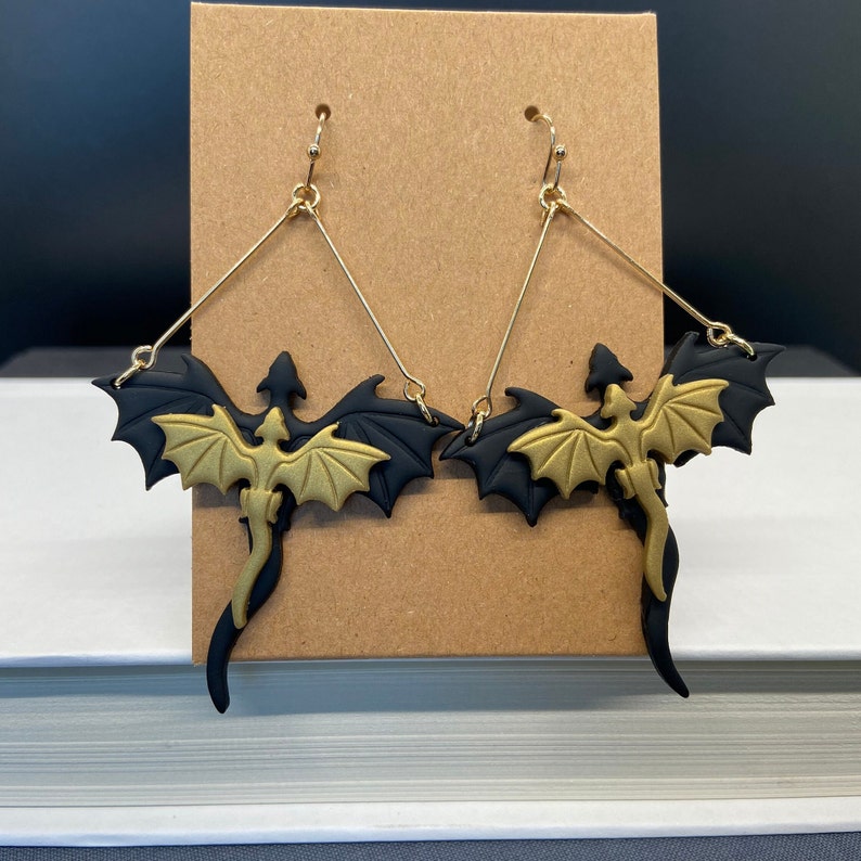 May include: A pair of gold and black dragon earrings. The dragons are in a flying pose with their wings spread out. The earrings are made of polymer clay and have gold hooks.
