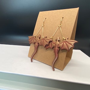 May include: A pair of gold dangle earrings with a red-brown dragon design. The dragons have wings and tails and are suspended from the earrings by a gold wire.
