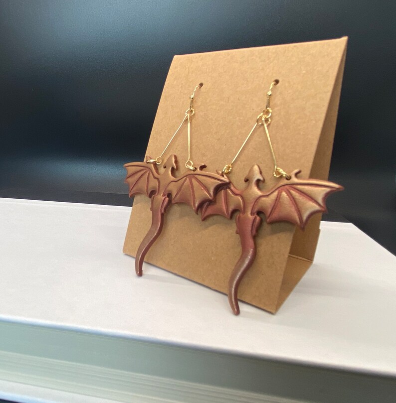 May include: A pair of gold dangle earrings with a red-brown dragon design. The dragons have wings and tails and are suspended from the earrings by a gold wire.