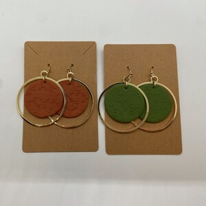 May include: Two pairs of gold hoop earrings with a round, patterned disc in the center. One pair has a brown disc with a floral pattern, and the other pair has a green disc with a floral pattern.