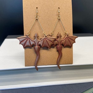 May include: A pair of gold dangle earrings featuring a brown dragon design. The dragons have wings and tails and are made of polymer clay.
