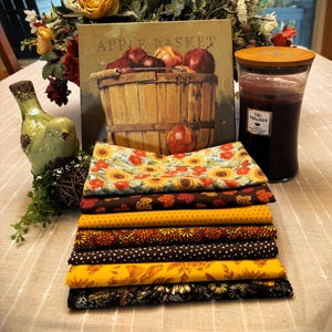 May include: A stack of seven folded fabric squares featuring autumn-themed patterns including sunflowers, maple leaves, polka dots, and floral motifs in shades of yellow, orange, brown, and black, arranged in a neat pile on a textured surface.