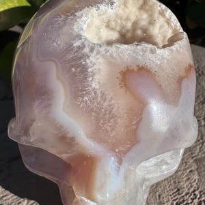 Puede incluir: Un cr&aacute;neo de &aacute;gata blanco y rosa con una gran cavidad de geoda de cristal blanco abierta. El cr&aacute;neo est&aacute; tallado en una piedra de &aacute;gata natural y tiene un acabado liso y pulido.