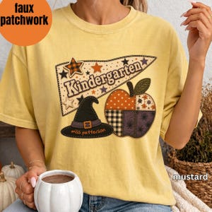 May include: A mustard yellow t-shirt featuring a graphic print of a pennant flag with the text Kindergarten, a black witch hat with the text miss patterson, and a patchwork-style apple. The design includes orange, black, and white patterns.