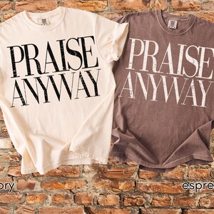 May include: Two short-sleeved t-shirts, one ivory and one espresso, are displayed. Each shirt has the words "PRAISE ANYWAY" printed in large, bold, black and white letters. The shirts are folded and placed on a brick background.