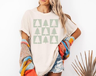 May include: Cream-colored t-shirt featuring a grid of nine Christmas trees in a light green hue. The trees are in a distressed style. The person is wearing a colorful sweater.
