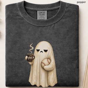 May include: A charcoal gray crew neck t-shirt featuring a graphic of a cream-colored ghost holding a brown mug with the word ghost printed on it and a baseball. The shirt has a Comfort Colors label at the neck and the color is labeled as pepper.