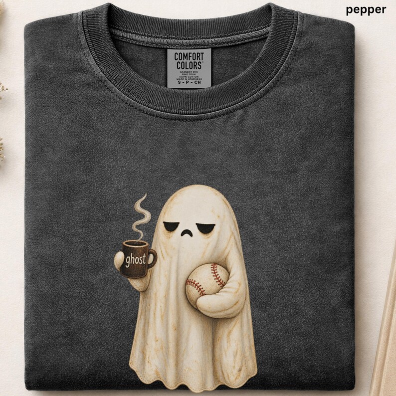 May include: A charcoal gray crew neck t-shirt featuring a graphic of a cream-colored ghost holding a brown mug with the word ghost printed on it and a baseball. The shirt has a Comfort Colors label at the neck and the color is labeled as pepper.