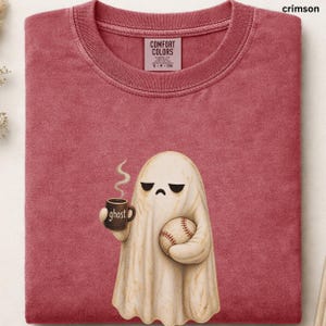 May include: A crimson crew neck t-shirt featuring a graphic of a cream-colored ghost with a grumpy expression. The ghost holds a brown mug with the word ghost printed on it and a baseball. The shirt has a Comfort Colors brand label inside.