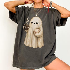 May include: A charcoal gray t-shirt featuring a graphic of a cream-colored ghost with a grumpy expression. The ghost holds a dark mug with the word ghost printed on it and a baseball. The shirt has short sleeves and a crew neckline.