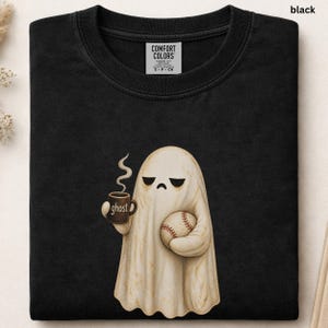 May include: A black crew neck t-shirt featuring a graphic of a beige ghost with a sad expression holding a steaming brown mug labeled ghost and a white baseball with red stitching. The shirt has a Comfort Colors brand label at the neckline.