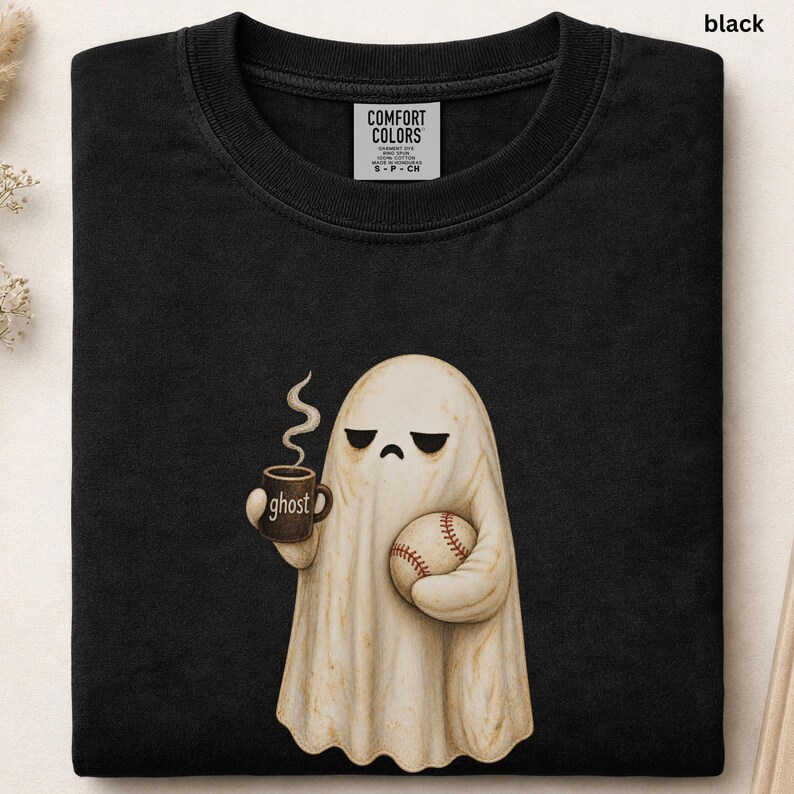 May include: A black crew neck t-shirt featuring a graphic of a beige ghost with a sad expression holding a steaming brown mug labeled ghost and a white baseball with red stitching. The shirt has a Comfort Colors brand label at the neckline.