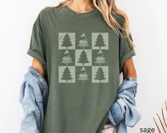 May include: Sage green t-shirt featuring a grid of nine Christmas tree graphics in varying shades of green. The trees are set within light green squares, creating a repeating pattern. The shirt has short sleeves and a relaxed fit.