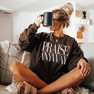 May include: A dark brown sweatshirt with the words "PRAISE ANYWAY" in white lettering. The person is holding a black mug. The person is wearing rings and a necklace. The person is wearing fuzzy slippers.