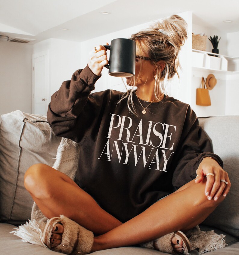 May include: A dark brown sweatshirt with the words "PRAISE ANYWAY" in white lettering. The person is holding a black mug. The person is wearing rings and a necklace. The person is wearing fuzzy slippers.