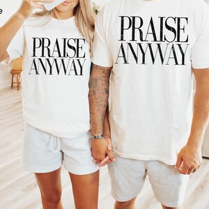 May include: White t-shirts with the text "PRAISE ANYWAY" in black, worn with gray shorts. The image shows two people holding hands, wearing matching outfits. The word "white" is in the top left corner.