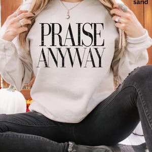 May include: A sand-colored sweatshirt with the words "PRAISE ANYWAY" in bold, black letters. The sweatshirt is a neutral color and has long sleeves. The person is wearing black jeans.