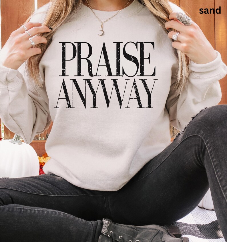 May include: A sand-colored sweatshirt with the words "PRAISE ANYWAY" in bold, black letters. The sweatshirt is a neutral color and has long sleeves. The person is wearing black jeans.