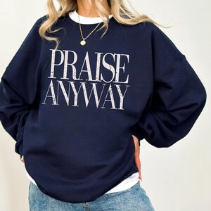 Distressed typography design reading praise anyway printed on a navy sweatshirt, styled as a faith-based inspirational garment for casual wear or church use