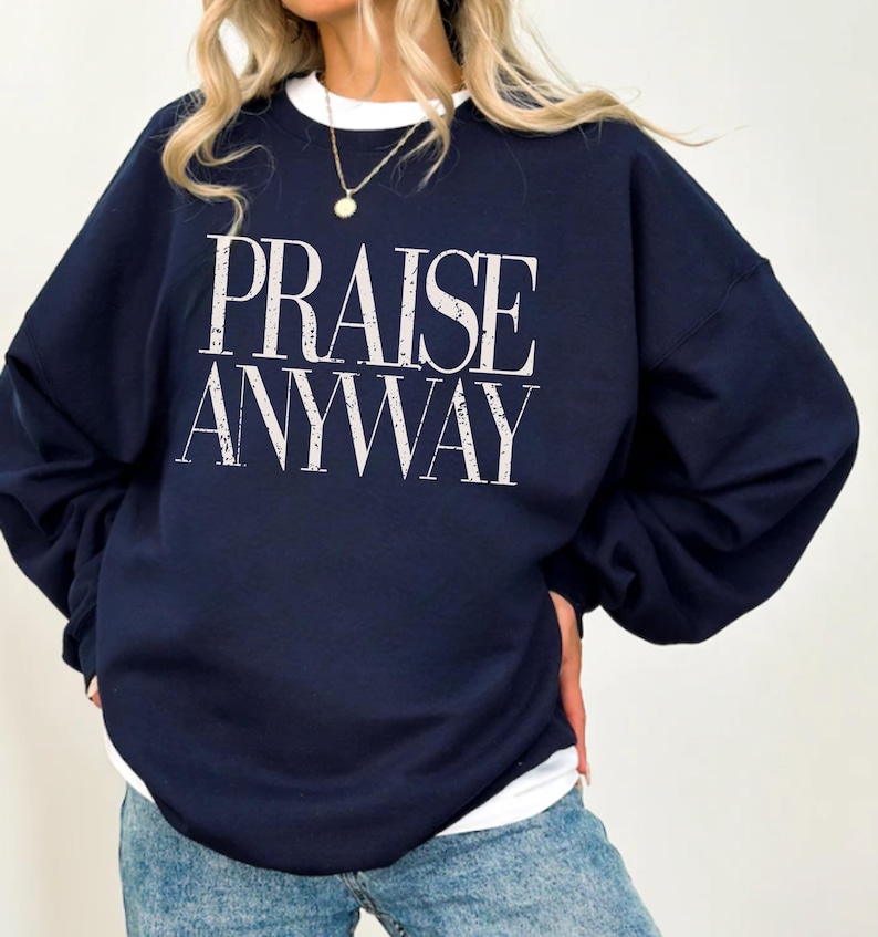 Distressed typography design reading praise anyway printed on a navy sweatshirt, styled as a faith-based inspirational garment for casual wear or church use