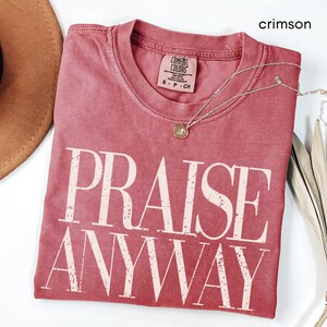 May include: Crimson-colored t-shirt with the words "PRAISE ANYWAY" in large, off-white block letters. The shirt has a crew neck and a small tag. A delicate silver necklace with a pendant is draped across the shirt. A brown hat and dried plant are also in the image.