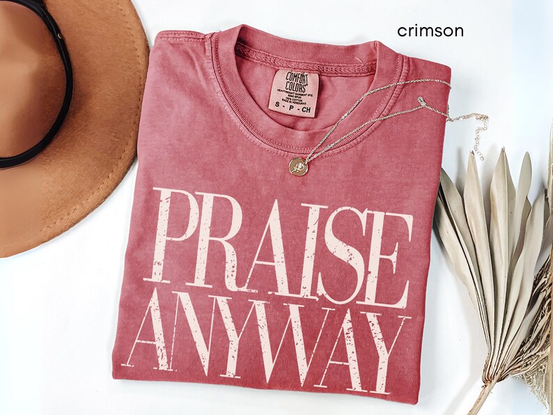 May include: Crimson-colored t-shirt with the words "PRAISE ANYWAY" in large, off-white block letters. The shirt has a crew neck and a small tag. A delicate silver necklace with a pendant is draped across the shirt. A brown hat and dried plant are also in the image.