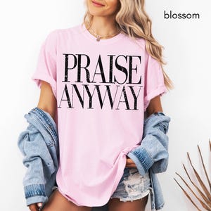 May include: A light pink t-shirt with the words "PRAISE ANYWAY" printed in black. The shirt is worn with a denim jacket and shorts. The word "blossom" is written in black in the upper right corner.
