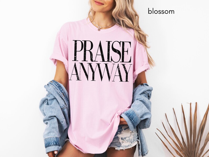 May include: A light pink t-shirt with the words "PRAISE ANYWAY" printed in black. The shirt is worn with a denim jacket and shorts. The word "blossom" is written in black in the upper right corner.