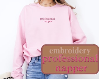 May include: A light pink sweatshirt with the embroidered text "professional napper" in a darker pink font. The image also shows a brown rectangle with the text "embroidery professional napper". The model is wearing blue jeans.