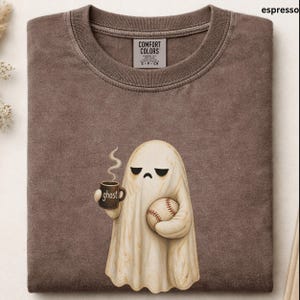 May include: A brown crewneck t-shirt featuring a graphic of a cream-colored ghost with a sad expression. The ghost holds a brown mug with the text ghost printed on it and a baseball. The shirt has a Comfort Colors label inside the collar.
