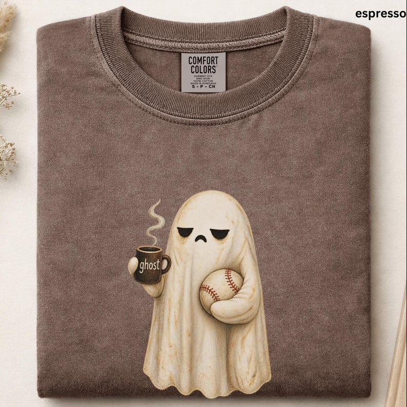 May include: A brown crewneck t-shirt featuring a graphic of a cream-colored ghost with a sad expression. The ghost holds a brown mug with the text ghost printed on it and a baseball. The shirt has a Comfort Colors label inside the collar.