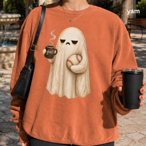 May include: A burnt orange crewneck sweatshirt featuring a graphic of a cream-colored ghost with a grumpy expression, holding a brown mug labeled ghost and a baseball. The sweatshirt has long sleeves and a ribbed neckline, worn with a black shoulder bag.