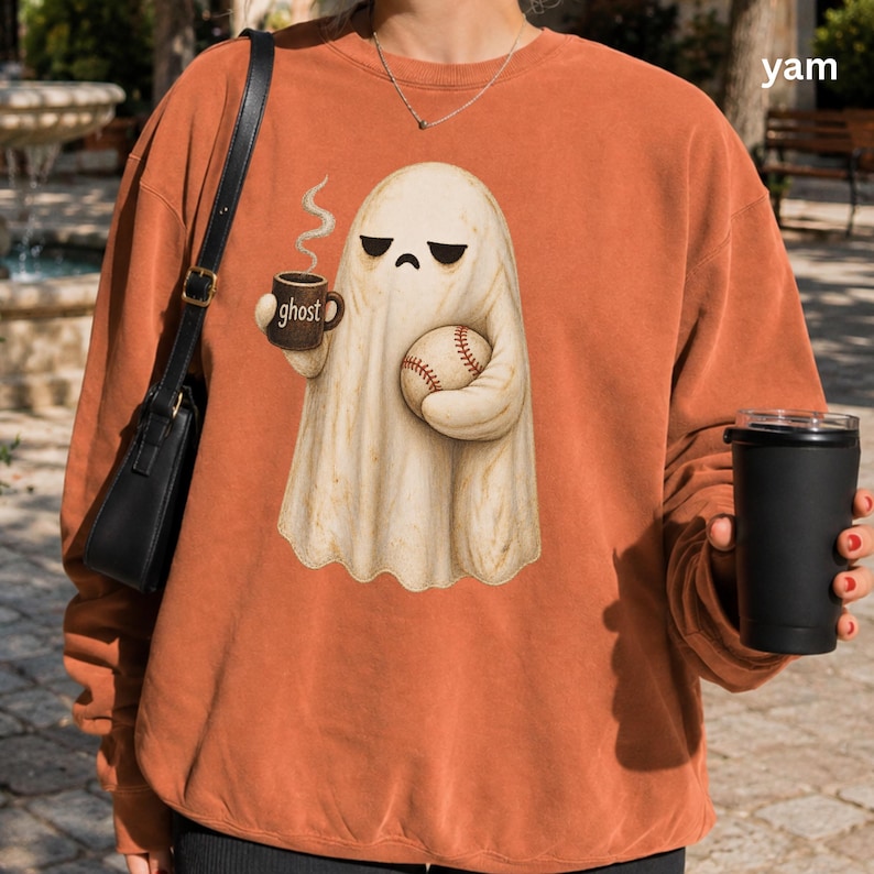 May include: A burnt orange crewneck sweatshirt featuring a graphic of a cream-colored ghost with a grumpy expression, holding a brown mug labeled ghost and a baseball. The sweatshirt has long sleeves and a ribbed neckline, worn with a black shoulder bag.
