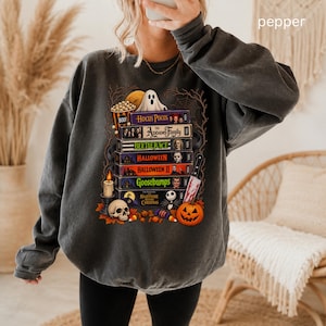 May include: A charcoal gray crewneck sweatshirt featuring a graphic print of stacked VHS tapes labeled with horror movie titles including Hocus Pocus, The Addams Family, Beetlejuice, Halloween, and Goosebumps, alongside a ghost, skull, and pumpkin.